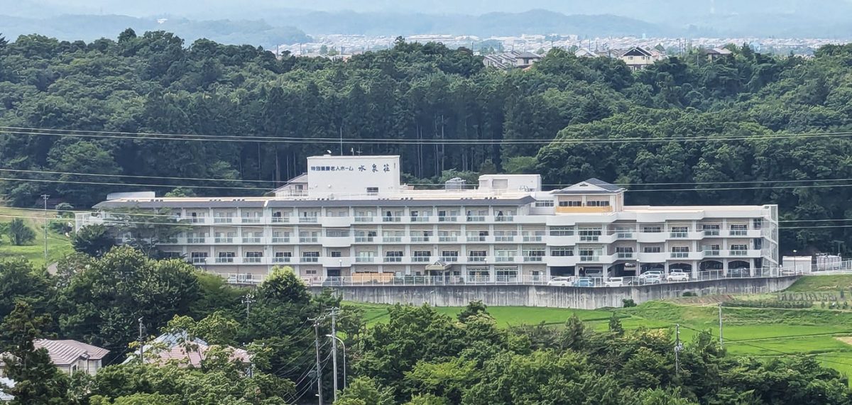 緑に囲まれた介護施設の全景。白い大規模建物が山間部に位置する。